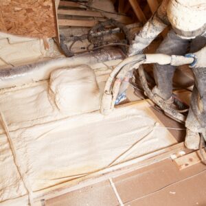 A professional installer in a protective helmet applies a thick layer of spray foam insulation to the floor of an attic space.