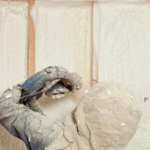 A close-up, over-the-shoulder view of a worker in dusty gloves holding a spray foam application gun.